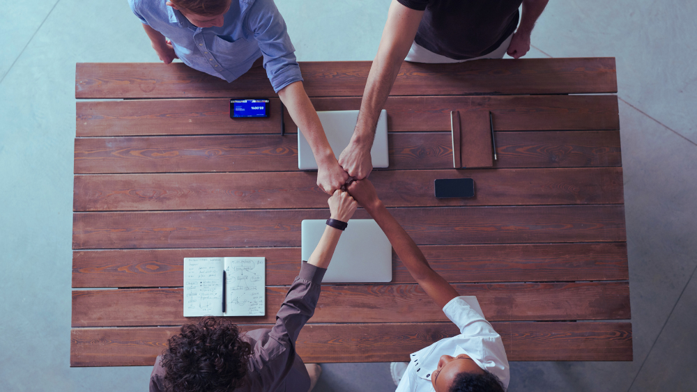 Ein Bild zeigt vier Personen, die an einem Holztisch sitzen und ihre Hände in der Mitte zu einem Handschlag vereinen, symbolisiert Zusammenarbeit und Fairness.